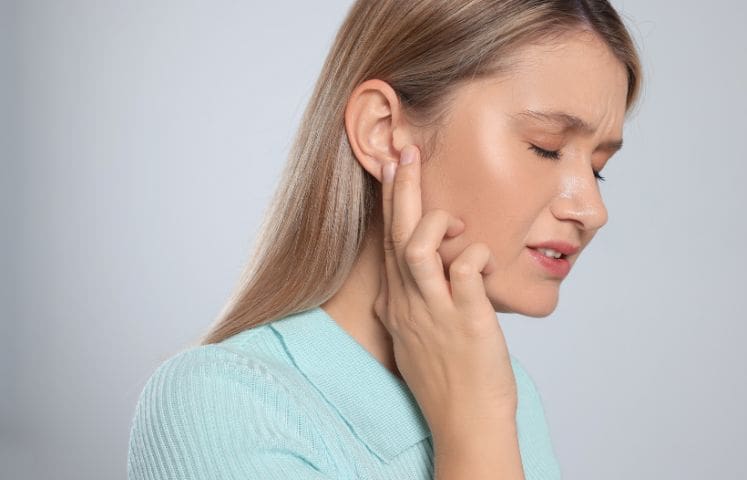 Person with long, straight, light brown hair, wearing a light blue sweater, holding a hand to the ear as if listening or indicating ear pain. The background is plain and light-colored.