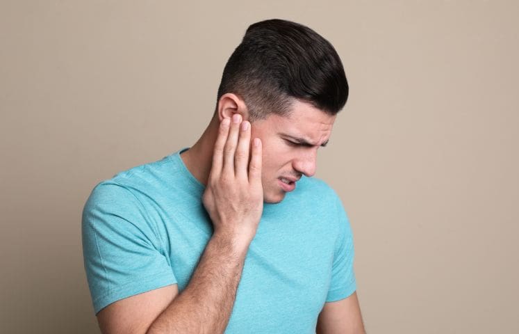 A person wearing a light blue t-shirt holding the left side of their head with their left hand. The face is blurred, and the background is beige. The image appears related to health, specifically ear pain or headaches