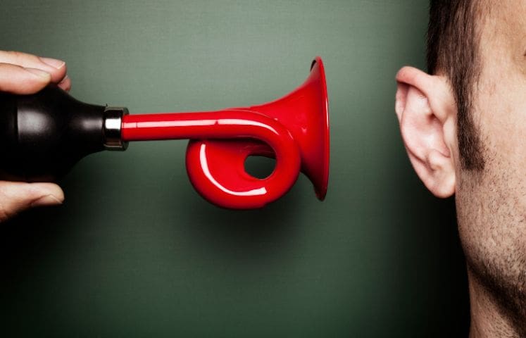 A person holds a red air horn close to another person's ear, suggesting the imminent use of the air horn, which could result in a loud noise. The scene captures a potentially humorous or startling moment, emphasizing the contrast between the quiet ear and the loud air horn.