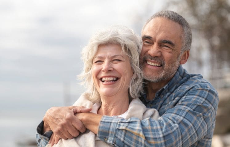 An elderly couple enjoying an affectionate moment outdoors. The man, in a blue plaid shirt, embraces the woman, who is draped in a light-colored shawl. The background features a natural setting with trees, water, and a cloudy sky.