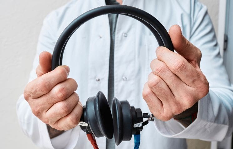Healthcare professional in a white coat holding a pair of black audiometry headphones.