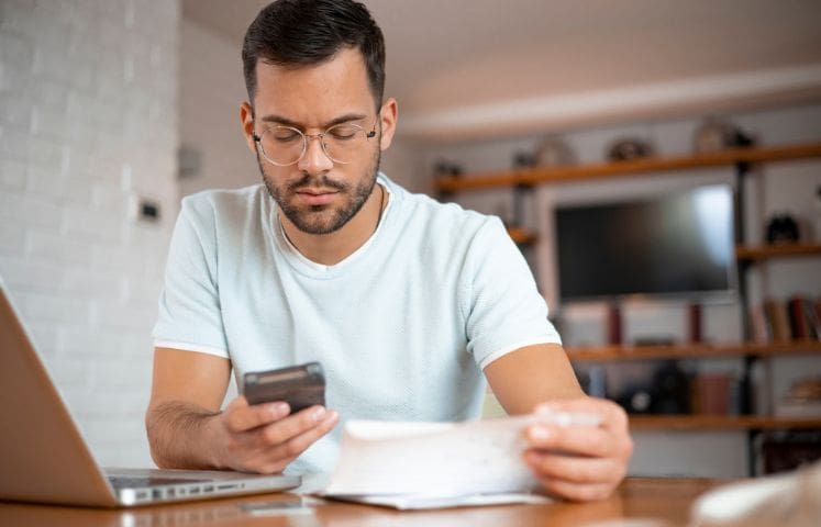 Man sitting at a table looking at his smartphone while holding a paper, with a laptop open in front of him.
