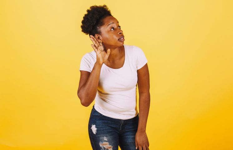 Young woman cupping her ear and leaning forward, indicating difficulty hearing, against a bright yellow background.