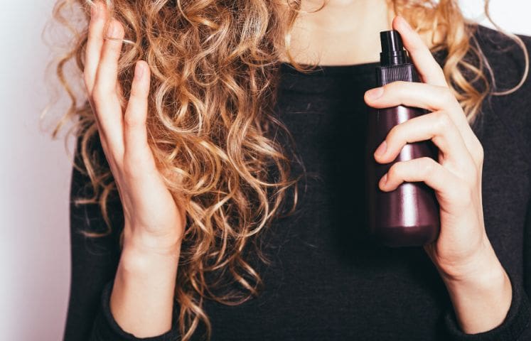 Woman with curly hair holding a spray bottle near her head, possibly applying hair product.